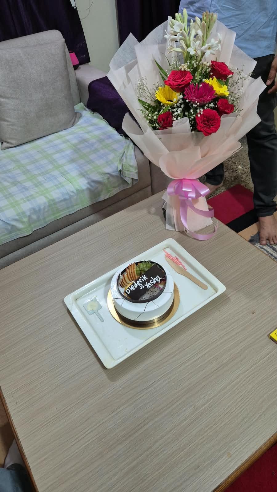 A cake reading "Diederik & Yasha" on a coffee table, with a big bouquet of roses, gerberas and sunflowers behind it